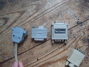 Three different DB15 to VGA adapters placed on a desk. From left to right; the first one is a custom made one, with both connectors seperated at different ends on a short cable. Then there's a Lindy adapter, which is more compact with both connectors together (DB15 on one end and VGA on the other) and there's a Lindy branding sticker added to the adapter itself. Finally, the one the right is unbranded with a similar compact all-in-one design, but includes a set of ten dipswitches for configuring the video mode you want to use.