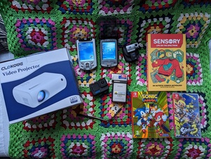 A selection of gifts I got placed on a crochet blanket. From the left; there's a Clokowe Video Projector in the box, two HP iPAQ Pocket PCs (the h5550 and hx2490b respectively), a HP Photosmart Mobile Camera that uses the SDIO interface, GoPro Hero 10 Black, Sony HDR-TG3E, a comic book called Sensory: Life on the Spectrum, along with Sonic Mania Plus and Splatoon 3 for the Nintendo Switch.
