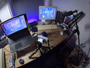 Filming setup I used for the bonus segment in the LC III video, showing an iMac G4 at the corner of a desk. A Sony a5100 camera is placed near the edge of the desk with a Joby Gorillapod Tripod, filming the keyboard and mouse connected to the iMac. Above the desk slightly is a Deity V-Mic D3 microphone on a stand, which is facing down towards the iMac for recording audio for the video. That microphone is connected with a long cable to the Zoom H1 audio recorder, placed next to the camera in this photo. To the left of the iMac is a Lenovo laptop running Linux, which was recording VGA capture footage from the iMac G4 using an Avermedia HDMI capture card and an Atlona scaler (not pictured). The laptop is displaying OBS with the live preview from the capture card, along with an mpv window playing back the capture I recorded previously