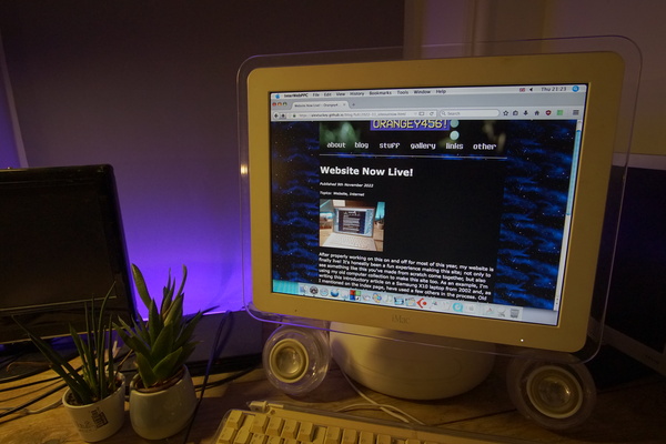 Picture of my iMac G4 setup in the corner of a desk with purple strip lighting behind it, and two IKEA desk plants to the left of it. The iMac is displaying the first article from my website, using the InterWebPPC web browser under Mac OS X Tiger.
