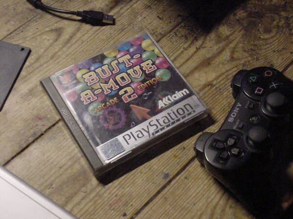 A physical PAL copy of Bust-A-Move 2 Arcade Edition for the PlayStation 1 placed on a desk, taken with the Mavica from a tripod. A DualShock 3 controller (for the PS3) can also be seen in the right corner, along with a floppy disk in the left corner