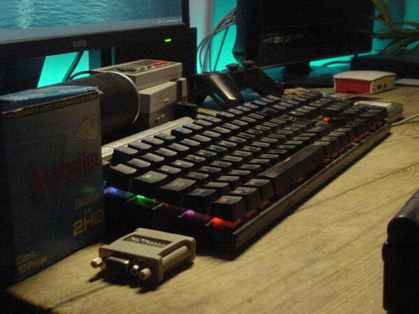 A side profile shot of a desktop PC setup, showing an Aukey KM-G6 mechanical keyboard with a 6-colour backlight, connected to an old HP Compaq Core 2 Duo desktop (off-screen). The monitor connected to it (a modern BenQ one) can be seen in the top left, along with an NES Classic Mini, a box of Verbatim floppy disks and a DB15 to VGA adapter (for old Macs). There's also a Raspberry Pi 3 and another generic HDTV in the top right, and the back of the desk has an LED strip light, which was set to a green colour here.