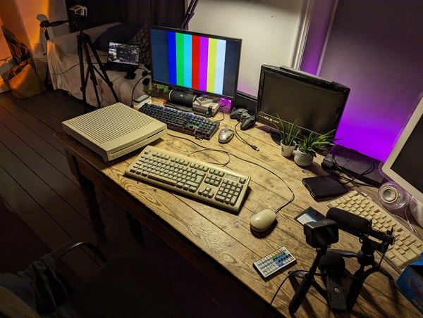 Filming setup I used for the main segment in the LC III video, focusing on that computer placed at the far left corner of a desk. There's also an Apple Extended Keyboard II and Desktop Bus Mouse II connected to the LC III. To the right of the photo is where the main camera equipment was, with a GoPro Hero 10 Black and Deity V-Mic D3 microphone both on small tripods, along with a Zoom H1 audio recorder that the Deity mic was connected to. A BenQ 22 inch 1080p monitor is shown to the right of the LC III and was displaying a colour bar output from the video scaler I used with the LC III (not pictured). Finally in the background, there's a sofa with my 2017 MacBook Pro I used for recording the VGA capture footage, along with a Sony a5100 camera on a standard tripod